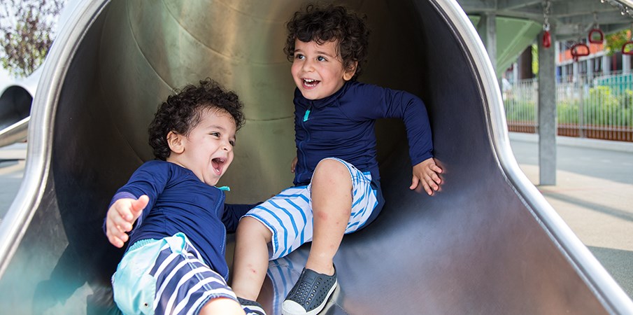 年轻的双胞胎男孩在Domino公园滑下一处不锈钢操场时微笑。